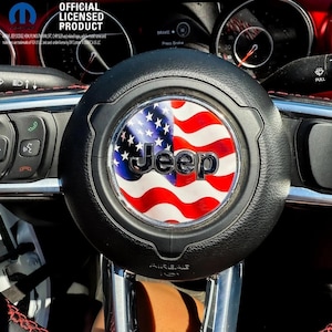 May include: A black leather Jeep steering wheel featuring a circular emblem with a waving American flag design and the silver Jeep logo in the center. The steering wheel includes integrated control buttons and is positioned in front of a vehicle dashboard.
