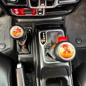 May include: A vehicle centre console featuring a gear shifter and a transfer case lever, both topped with circular inserts displaying a sunset palm tree graphic. The black leather boots have red stitching, and the console includes a PRND gear indicator.