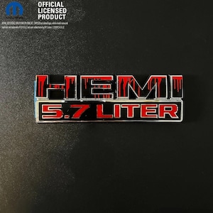 May include: A rectangular vehicle emblem featuring the word HEMI in black with red drip-style accents, positioned above the text 5.7 LITER in red. The emblem includes a Mopar logo and the text OFFICIAL LICENSED PRODUCT in the upper left corner.