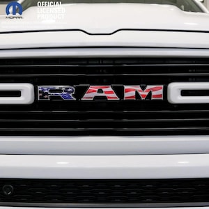 May include: Close-up of a white truck grille with the word "RAM" in the center, featuring a patriotic American flag design. The grille is black with white accents. The top left corner has the Mopar logo and the text "OFFICIAL LICENSED PRODUCT."