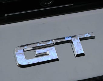 May include: A metallic GT emblem featuring a reflective chrome finish with a blue and white marbled pattern, mounted on a smooth, glossy black surface. The letters are stylized with sharp, angular edges and a three-dimensional appearance.