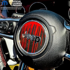 Peut inclure: Un emblème central de volant noir arborant un logo Jeep argenté sur un motif de gouttes verticales rouges et noires. L'emblème est entouré d'un anneau chromé et monté sur un volant noir texturé avec des surpiqûres rouges.