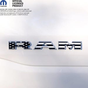 May include: A silver RAM emblem is displayed on a white vehicle. The letter "R" has a black and white American flag design with stars. The letters "A" and "M" have black and white stripes. The image also includes the MOPAR logo and the text "OFFICIAL LICENSED PRODUCT".