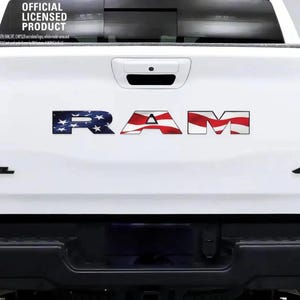 Può includere: Un portellone posteriore di un camion RAM bianco con una decalcomania con il motivo della bandiera americana applicato sulle lettere. Il logo Mopar e il testo Official Licensed Product appaiono in alto a sinistra, con un emblema Rebel nero e un badge 4x4 visibili.