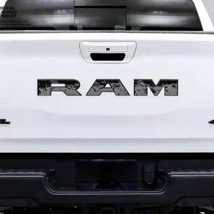 May include: White truck tailgate featuring the black letters "RAM" across the centre. The word "REBEL" is on the left, and "4x4" on the right. The letters have a distressed, textured appearance.