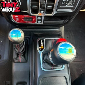 May include: Car interior with custom shift knobs displaying beach scenes. The shift knobs have a black base with a circular image of a beach, palm tree, and blue sky. The image also shows the text "TINY WRAPZ".