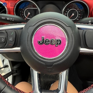 May include: Close-up of a black steering wheel with a pink Jeep logo in the center. The steering wheel has red stitching and is surrounded by the car's dashboard, including gauges and controls. The text "OFFICIAL LICENSED PRODUCT" is visible.