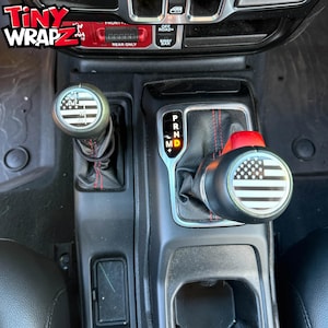 Peut inclure: Intérieur de véhicule avec des pommeaux de levier de vitesses personnalisés. Les pommeaux noirs arborent un motif drapeau américain noir et blanc. Le tableau de bord présente également des détails rouges et le texte "Tiny Wrapz".