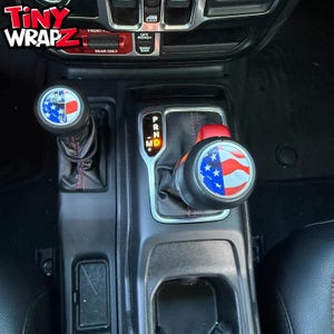 Peut inclure: Vue intérieure d'une console de véhicule avec des pommeaux de levier de vitesses personnalisés. Les pommeaux affichent un motif patriotique avec le drapeau américain. Le texte "Tiny Wrapz" est visible en haut à gauche.