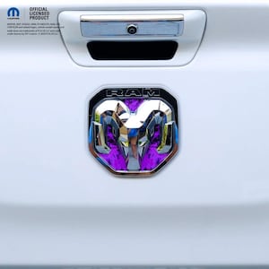 May include: A white vehicle tailgate featuring a chrome RAM emblem with a purple reflective insert. Above the emblem is a chrome handle with a built-in camera. Text in the upper left corner reads MOPAR OFFICIAL LICENSED PRODUCT and legal text.