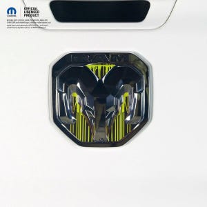 May include: A chrome Ram truck emblem featuring a stylized ram head logo with a bright neon yellow paint drip design behind the metallic surface. The emblem includes the word RAM at the top and is displayed on a white surface with official Mopar text.