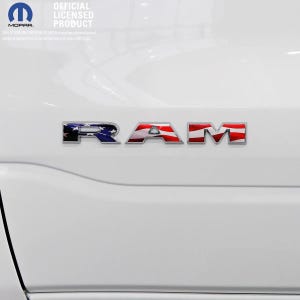 May include: A close-up of a white vehicle's body with the chrome letters "RAM" in the center. The letters are designed with a patriotic American flag theme. The image also includes the Mopar logo and the text "OFFICIAL LICENSED PRODUCT".