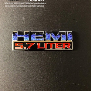 May include: A rectangular metal emblem with the text "HEMI" in black letters with blue flame accents, above "5.7 LITER" in red. The emblem also includes the Mopar logo and the text "OFFICIAL LICENSED PRODUCT".
