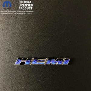 May include: A silver HEMI emblem featuring a blue and black flame pattern inlay. The text OFFICIAL LICENSED PRODUCT and the Mopar logo are printed in white at the top left corner, along with additional small legal text regarding trademark ownership.