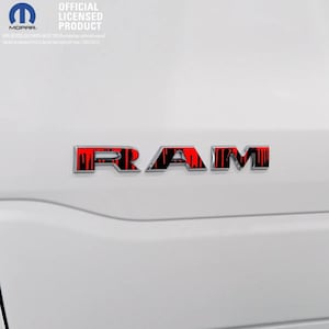 Peut inclure: Gros plan sur un véhicule blanc avec un emblème "RAM" rouge et noir. L'emblème présente un design unique, et le véhicule affiche également le logo "MOPAR" et le texte "OFFICIAL LICENSED PRODUCT".