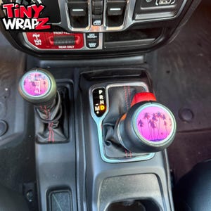 Könnte beinhalten: Schwarze und silberne Schalthebelknöpfe für Autos mit einem rosa-violetten Palmenmuster. Die Knöpfe sind Teil eines Autoinnenraums. Das Bild zeigt auch den Text "Tiny Wrapz".