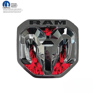 May include: A RAM vehicle emblem featuring a chrome-finished ram head logo centered over a vibrant red flame pattern background. The word RAM is embossed in black lettering at the top of the shield-shaped badge, which includes official Mopar licensing text.