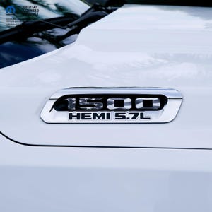 May include: Close-up of a white vehicle hood with a chrome emblem. The emblem reads "1500 HEMI 5.7L" in black lettering. The "1500" has a flag design. The vehicle is an official licensed product.