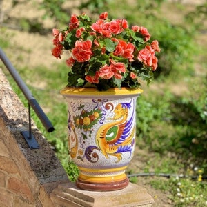 Könnte beinhalten: Ein weißer Keramiktopf mit einem bunten Blumen- und Vogeldesign. Der Topf ist mit rosa Blüten und grünen Blättern gefüllt.
