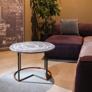 May include: A round, grey coffee table with a white abstract design on the tabletop. The table has a dark metal frame with a circular base and four legs. The table is approximately 61 cm in diameter.
