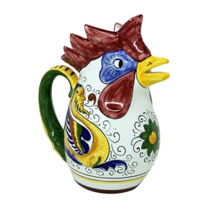 May include: A ceramic rooster-shaped pitcher. The pitcher has a white body with a rooster head design in red, blue, and yellow. The handle is green and yellow, and it is decorated with floral and scroll patterns. A decorative item.