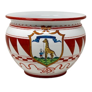 May include: White ceramic planter with a red rim and red and white geometric pattern. A shield with a giraffe and a person holding a rope is painted on the front. The shield has a blue ribbon with the text "Giraffeville I. Scout".