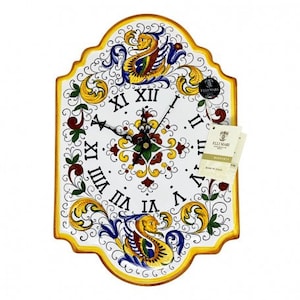 May include: A white ceramic clock with a yellow, blue, and red floral design. The clock face has Roman numerals and a black clock hand. The clock is made in Italy by the brand "Felli Mari".