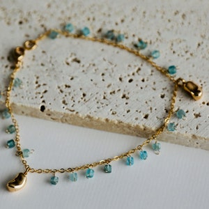 Op de afbeelding: Een gouden kettingarmband met blauwe edelsteenbedels. De armband heeft twee gouden bedels, een in de vorm van een boon en de andere in de vorm van een voet.