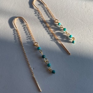 May include: A pair of gold chain earrings with turquoise beads dangling from the bottom. The earrings are on a white surface.