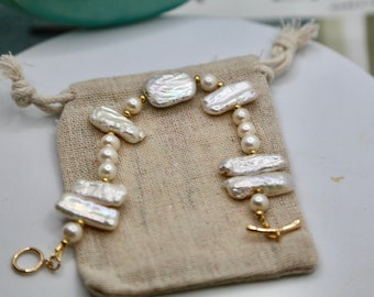 May include: A white pearl bracelet with gold accents. The bracelet is made of irregular shaped pearls and small round pearls. The bracelet is on a beige fabric background.