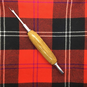 May include: A wooden handled kilt pin with a metal hook and a silver tip. The pin is lying on a red, black, and white plaid fabric.