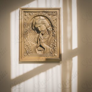 May include: A wooden framed picture of a carved image of Jesus with his hands clasped in prayer. The image is in a light brown color and is set against a white wall.