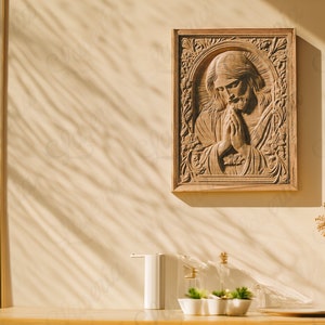 May include: A wooden wall decor featuring a carved image of Jesus praying with his hands clasped together. The decor is framed in a light brown wood frame and hangs on a beige wall. The wall has a shadow pattern from sunlight shining through a window.