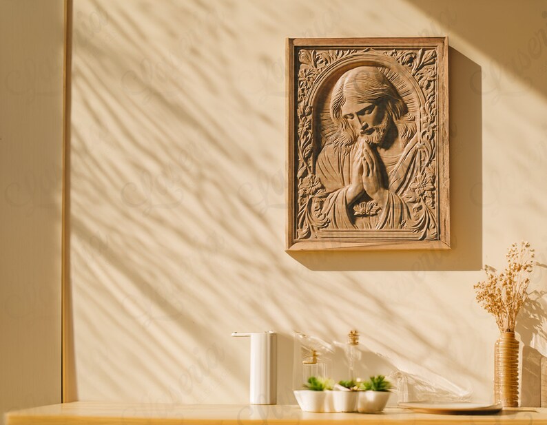 May include: A wooden wall decor featuring a carved image of Jesus praying with his hands clasped together. The decor is framed in a light brown wood frame and hangs on a beige wall. The wall has a shadow pattern from sunlight shining through a window.