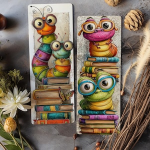 May include: Two whimsical bookmarks featuring cartoon caterpillars. One bookmark shows two caterpillars atop a stack of books, with a butterfly. The other bookmark shows two caterpillars wearing glasses, also atop stacks of books. The bookmarks have a white background.