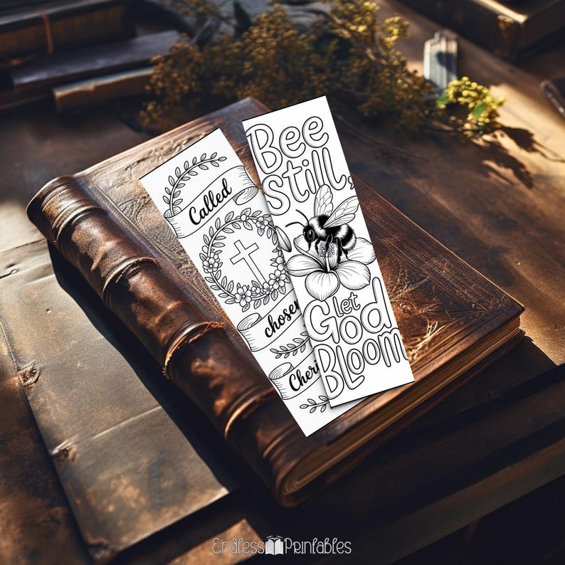 Pode incluir: Dois marcadores brancos com texto e ilustra&ccedil;&otilde;es pretas repousam sobre um livro antigo encadernado em couro. Um marcador diz "Called, chosen, Cherished" com uma cruz e uma coroa floral. O outro diz "Bee Still, Let God Bloom" com uma abelha em uma flor.