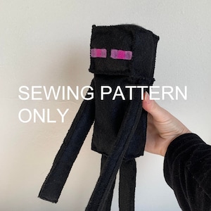 May include: Sewing pattern for a black felt Minecraft Creeper plush toy. The Creeper has pink eyes and is standing on a white surface.