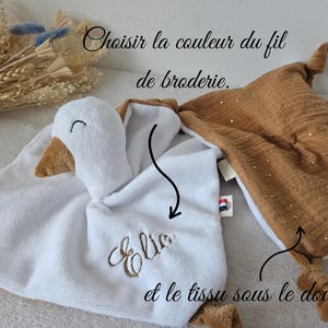 May include: A white and brown plush baby comforter with a goose head and embroidered name. A second brown comforter with gold accents is also shown. Text in French reads "Choose the color of the embroidery thread and the fabric under the comforter."