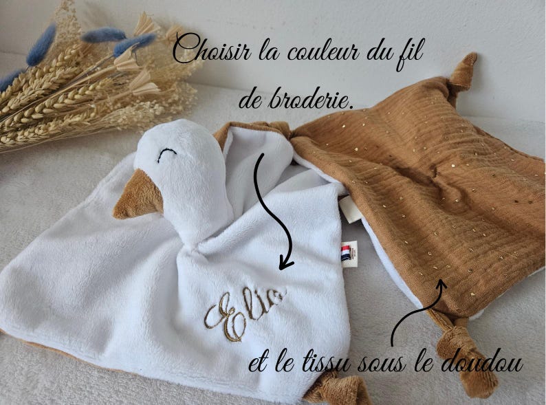 May include: A white and brown plush baby comforter with a goose head and embroidered name. A second brown comforter with gold accents is also shown. Text in French reads "Choose the color of the embroidery thread and the fabric under the comforter."