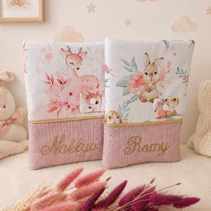 Pu&ograve; includere: Due copertine rettangolari in tessuto con parti superiori bianche decorate da illustrazioni ad acquerello di un cerbiatto e un coniglietto circondati da fiori. Le basi sono rosa polvere con bordo dorato e i nomi Na&eacute;lya e Romy ricamati.