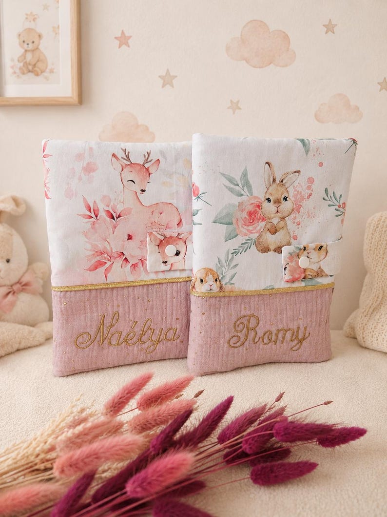 Pu&ograve; includere: Due copertine rettangolari in tessuto con parti superiori bianche decorate da illustrazioni ad acquerello di un cerbiatto e un coniglietto circondati da fiori. Le basi sono rosa polvere con bordo dorato e i nomi Na&eacute;lya e Romy ricamati.