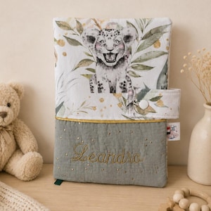 Pu&ograve; includere: Una copertina per libro rettangolare in tessuto con una sezione superiore bianca raffigurante un cucciolo di leopardo e fogliame verde, e una sezione inferiore trapuntata verde salvia con il nome Leandro ricamato in filo dorato e stelline.
