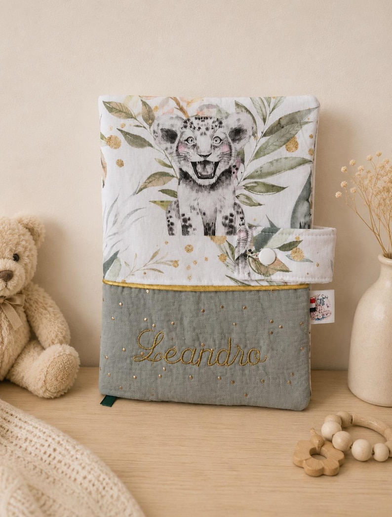 Pu&ograve; includere: Una copertina per libro rettangolare in tessuto con una sezione superiore bianca raffigurante un cucciolo di leopardo e fogliame verde, e una sezione inferiore trapuntata verde salvia con il nome Leandro ricamato in filo dorato e stelline.