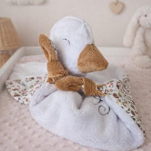May include: A white plush duck-shaped baby comforter with brown accents, a floral patterned edge, and a small brown bunny toy attached. The comforter is on a pink surface.