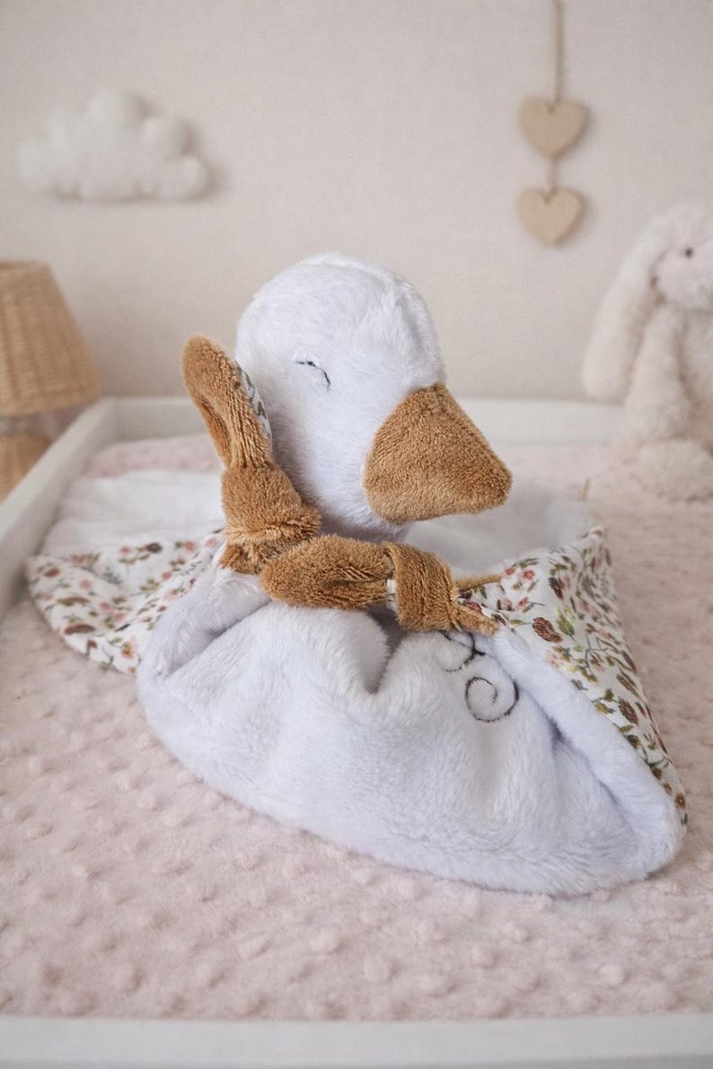 May include: A white plush duck-shaped baby comforter with brown accents, a floral patterned edge, and a small brown bunny toy attached. The comforter is on a pink surface.