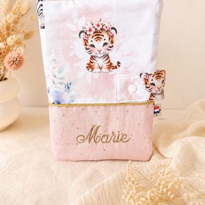 Pu&ograve; includere: Una custodia in tessuto rettangolare con una sezione superiore bianca raffigurante un cucciolo di tigre con una corona di fiori e una sezione inferiore rosa con il nome Marie ricamato in filo dorato, chiusa da un bottone a pressione bianco.