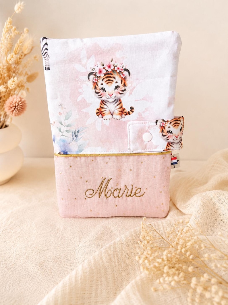 Pu&ograve; includere: Una custodia in tessuto rettangolare con una sezione superiore bianca raffigurante un cucciolo di tigre con una corona di fiori e una sezione inferiore rosa con il nome Marie ricamato in filo dorato, chiusa da un bottone a pressione bianco.