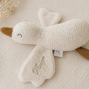 Peut inclure: Une peluche en forme d'oie de couleur crème en tissu sherpa texturé. L'oie présente un bec et des pattes marron, des yeux fermés brodés et le prénom "Hugo" cousu en écriture cursive sur une aile. Une petite étiquette blanche est fixée sur le côté.
