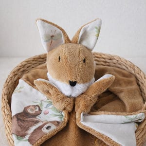 Peut inclure: Une peluche lapin marron avec de longues oreilles et un col blanc, dans un panier en osier. La couverture est ornée d'un tissu doux à motifs avec une illustration de paresseux et des accents de libellules, créant un design douillet et réconfortant.