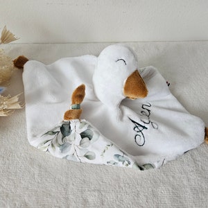 May include: A white plush baby comforter with a goose head and brown beak and feet. The comforter has a white and green floral patterned corner and the name "Agen" embroidered in black.